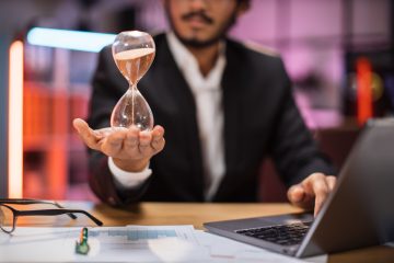 Focus on hand of smiling confident businessman in formal suit sitting at desk with modern laptop and holding hourglass. Concept of deadline people and office work.